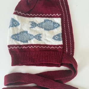 May include: A deep red knitted bonnet featuring a wide white horizontal band across the center. The white band is decorated with a repeating pattern of light blue fish. The bonnet includes long red knitted ties with white tips at the ends.