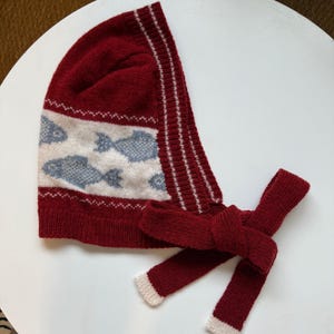 May include: Red knitted hat with a white and blue fish pattern and a long red tie. The hat has a white trim around the edge.