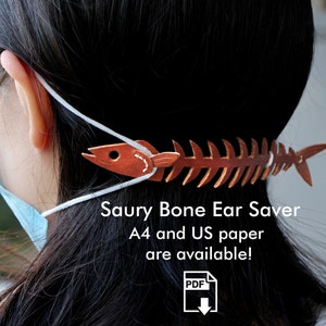 May include: A brown leather fishbone ear saver for masks. The ear saver is shaped like a fish skeleton with a fish head at one end. The text "Saury Bone Ear Saver" and "A4 and US paper are available!" is printed below the ear saver. A PDF download icon is below the text.