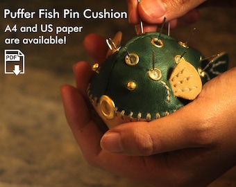 May include: A green leather pincushion shaped like a puffer fish with gold accents and a beige fin. The pincushion is being held in a hand with several pins sticking out of it.