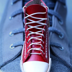 May include: A miniature red leather sneaker with white laces and a white toe cap. The sneaker is placed on top of a larger gray sneaker. The text "THE PARCHMENT HONGKONG Handcrafted Accessories" is visible.