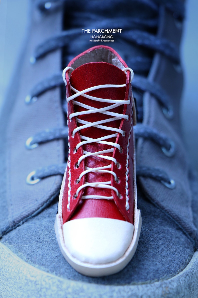 May include: A miniature red leather sneaker with white laces and a white toe cap. The sneaker is placed on top of a larger gray sneaker. The text "THE PARCHMENT HONGKONG Handcrafted Accessories" is visible.
