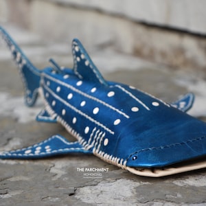 May include: A blue leather whale shark with white polka dots and stitching. The shark is lying on a grey textured surface. The text "THE PARCHMENT" and "HONOLULUGIRL" is visible on the surface.