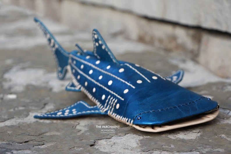 May include: A blue leather whale shark with white polka dots and stitching. The shark is lying on a grey textured surface. The text "THE PARCHMENT" and "HONOLULUGIRL" is visible on the surface.