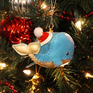 May include: A blue whale Christmas bauble with a white pom-pom hat and red Santa hat. The bauble is hanging on a green Christmas tree with red and gold baubles.