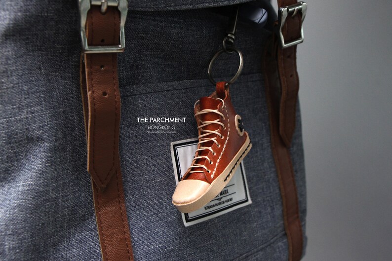 May include: A brown leather high-top sneaker keychain with white laces and a cream-colored toe. The keychain is attached to a gray backpack with brown leather straps. The text "THE PARCHMENT HONGKONG" is visible.