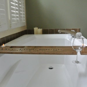 Puede incluir: Una bandeja de baño de madera con el texto "Live, Love, Laugh, and drink wine" grabado en ella. La bandeja tiene dos portavelas y un portavasos para vino. La bandeja está colocada sobre una bañera blanca.