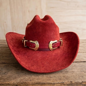 Könnte beinhalten: Ein roter Cowboyhut aus Wildleder mit breiter Krempe und eingedrückter Krone. Der Hut ist mit einem dunkelroten Lederband verziert, das zwei goldfarbene Metallschnallen mit aufwendigen Gravurdetails an der Vorderseite und den Seiten des Bandes enthält.