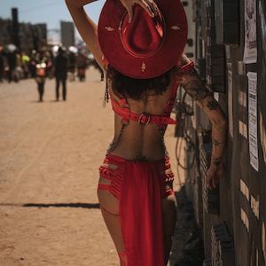 Könnte beinhalten: Eine Person trägt ein rotes Lederoutfit mit einem roten Cowboyhut und goldenen Akzenten. Die Person steht vor einer Wand, die mit Postern und anderen Gegenständen bedeckt ist.