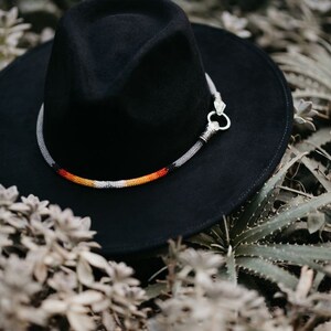 Puede incluir: Un sombrero fedora de fieltro negro con una banda de cuentas en tonos de blanco, naranja y rojo. El sombrero tiene una hebilla de metal plateado.