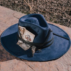 Peut inclure: Un chapeau fedora bleu marine à large bord. Le chapeau est orné d'une bande en cuir noir, d'une chaîne argentée et d'un pendentif en forme de crâne d'oiseau. Deux cartes de tarot sont glissées sous la bande. Les cartes ont des illustrations et le texte "PAGE of CUPS".