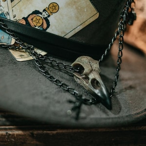 Puede incluir: Un collar de cadena negra con un colgante de cráneo de pájaro cuelga de un sombrero de fieltro marrón. El sombrero está decorado con cartas de tarot, incluida la carta 'IV', que presenta una figura sosteniendo un pentagrama.