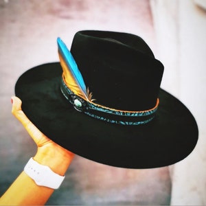 Puede incluir: Un sombrero fedora negro con ala ancha, adornado con una banda decorativa y una vibrante pluma azul y amarilla. El sombrero presenta un acento turquesa y un diseño clásico y elegante.