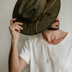 May include: A man wearing a green suede fedora with a brown leather band and a feather accent. The hat is covering his eyes and he is looking down.
