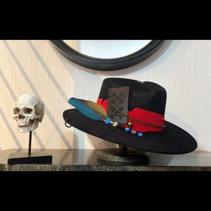 Op de afbeelding: Een zwarte cowboyhoed van vilt met een rode band, een veer en een ketting met een bel eraan. De hoed staat op een standaard en heeft een speelkaart in de band gestoken.