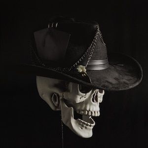 May include: A black cowboy hat with a silver chain and a small decorative skull is perched atop a white skull. The hat has a black tag attached. The image is set against a black background.