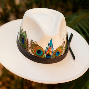 May include: A cream-coloured fedora hat with a wide brim. It has a black leather band and is decorated with peacock feather designs in green, blue, and brown. A colourful geometric design is centered above the brim.
