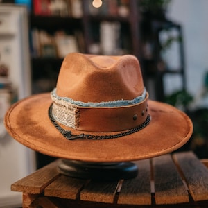 May include: A brown fedora hat with a wide brim. It has a denim band, a leather band with metal studs, and a black chain detail. The hat is displayed on a small wooden table.