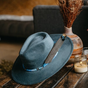 Könnte beinhalten: Ein schieferblauer Filzhut im Fedora-Stil mit einem Perlenhutband in Hellblau, Schwarz und Weiß. Eine einzelne Feder mit grau-braunem Muster ist in das Perlenband gesteckt und liegt seitlich am Hutkopf des Fedora-Hutes an.