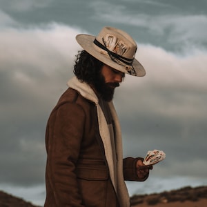 May include: A person wearing a brown suede jacket with a white fur collar and a white hat with a black band and a feather. The hat has a design of playing cards on it. The person is holding a deck of playing cards in their hand.