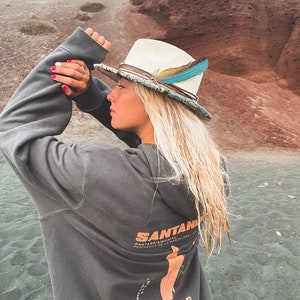 May include: A woman wearing a grey hoodie with a graphic print of a map and the text "SANTANDER CANTABRIA INFINITA PENINSULA DE LA MAGDALENA, CANTABRIA" and "YUXUS GRANADIRYT". She is wearing a white and blue hat with a feather.