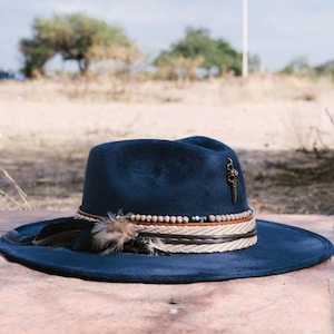 Könnte beinhalten: Ein tief dunkelblauer Filzhut im Fedora-Stil mit einem dekorativen Hutband aus beigefarbenem geflochtenem Seil, kleinen braunen Perlen und einem Lederriemen. Seitlich ist eine dunkle Feder sowie ein kleiner metallener Vogelschädel-Anhänger befestigt.