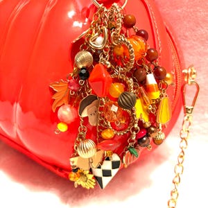 May include: A bright red, pumpkin-shaped handbag adorned with a large, multi-layered gold-tone charm cluster. The charms include miniature autumn-themed figures, colorful beads, a black and white checkered heart, and a gold chain shoulder strap.