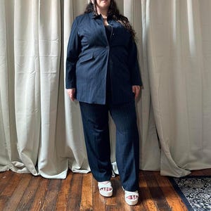 May include: A navy blue pinstripe suit, consisting of a blazer and wide-leg trousers. The blazer has a single button closure and front pockets. The person is wearing white platform sandals. The background features neutral-colored curtains and a wooden floor.