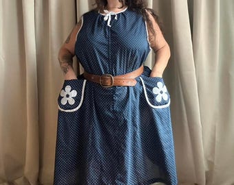 May include: A sleeveless navy blue dress with white polka dots and white trim. The dress features two front pockets with white flower appliqués and a brown belt. The dress is knee-length and has a tie at the neckline.