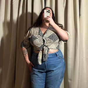 May include: A patterned, short-sleeved top with a tie front. The top features a mix of black and white geometric and paisley designs. The model is wearing blue jeans. The top is a neutral color, with a V-neckline.
