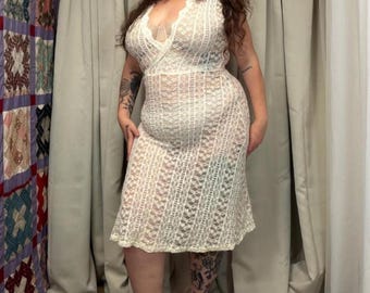 May include: A sleeveless midi-length dress made of cream-colored lace with a vertical striped pattern. The dress features a deep V-neckline with a wrap-style bodice, a fitted waist, and a flared skirt that falls below the knees in a semi-sheer fabric.