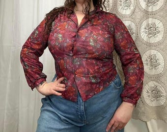 May include: A burgundy button-down shirt with a floral pattern in shades of red, orange, and green. The long-sleeved shirt is paired with light blue denim jeans.