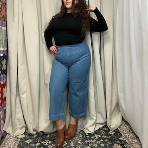 May include: A person wearing a black long-sleeve shirt and light blue denim pants. The pants are wide-legged and cropped. The person is also wearing brown leather clogs.