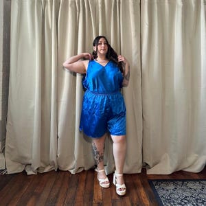 May include: A woman wearing a blue satin shorts set with a V-neck top. She is standing in front of a white curtain and wearing white sandals.