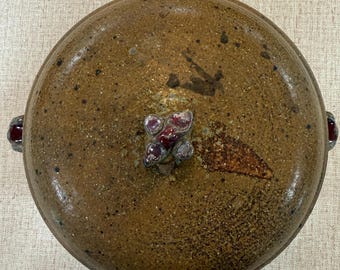May include: A round, brown ceramic casserole dish with a speckled glaze and a textured surface. The lid features a central knob with a dark red, glossy accent, and two small, matching red handles are visible on the sides of the vessel.