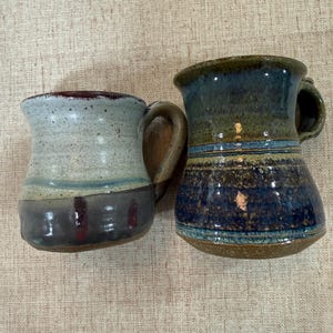 May include: Two ceramic mugs with handles. The left mug features a light gray upper section with a dark gray base and subtle red vertical accents. The right mug displays a mottled green and blue glaze with a textured, dark brown base finish.