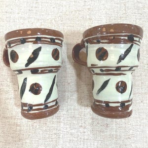 May include: Two ceramic mugs featuring a cream-colored base with hand-painted brown horizontal bands, dark brown circular spots, and elongated black brushstroke accents. Each mug has a sturdy brown handle and a wide, flared rim design.