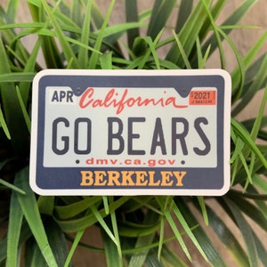 May include: A rectangular sticker resembling a California license plate. The sticker features the words "GO BEARS" in large, bold letters, with "California" and "BERKELEY" also printed on it. The sticker has a blue border and is set against a background of green foliage.