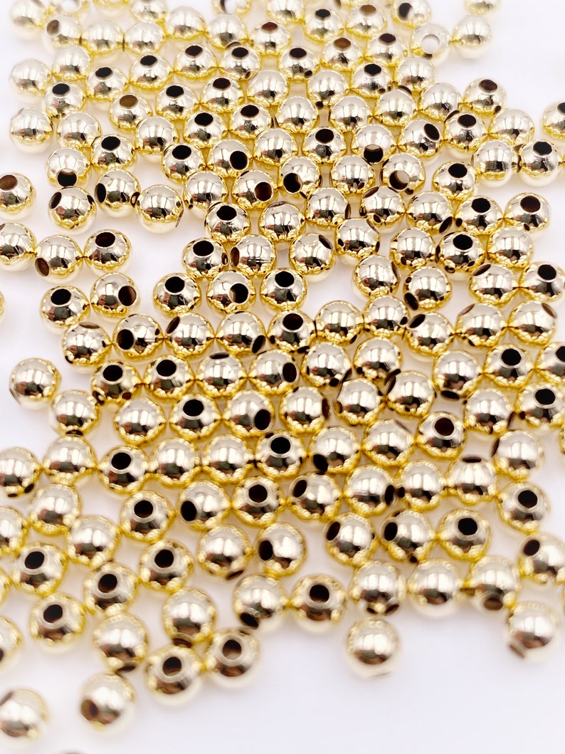 K&ouml;nnte beinhalten: Ein gro&szlig;er Haufen goldfarbener runder Perlen mit einem Loch in der Mitte. Die Perlen sind auf einer wei&szlig;en Oberfl&auml;che verstreut.