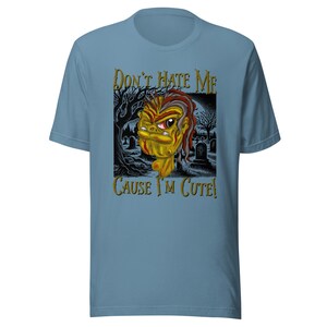 Könnte beinhalten: Hellblaues T-Shirt mit einem Cartoon-Motiv. Die Grafik zeigt ein gelbes, rotes und braunes Monster mit einer Friedhofsszene und dem Text "Don't Hate Me Cause I'm Cute!".
