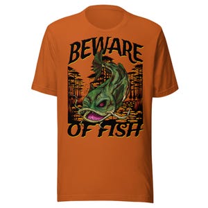 以下が含まれることがあります： オレンジ色のTシャツ。緑色のナマズのイラストと「BEWARE OF FISH」の文字が黒と黄色で描かれています。デザインには、木々と不気味な魚がいる沼地のシーンが含まれています。