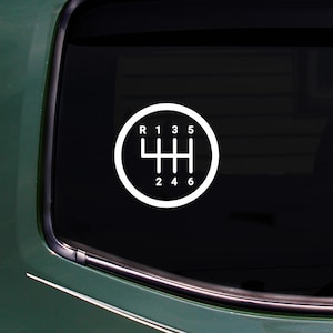 Może przedstawiać: Biała naklejka z wzorem zmiany biegów manualnej skrzyni biegów, pokazująca bieg wsteczny (R), biegi pierwszy (1), trzeci (3) i piąty (5) na górze oraz drugi (2), czwarty (4) i szósty (6) na dole. Projekt to okrąg z wzorem biegów w środku.