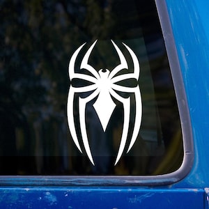 May include: White vinyl decal of a stylized spider emblem, featuring a central spider body with eight legs and a diamond-shaped element. The decal is displayed on a window, with a blue vehicle frame visible.