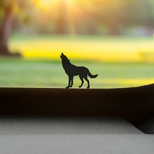 Könnte beinhalten: Eine schwarze Silhouette eines heulenden Wolfs vor einem verschwommenen Hintergrund aus grünem Gras und einem hellgelben Himmel. Der Wolf steht auf einer dunklen Oberfläche, mit der Sonne im Hintergrund.