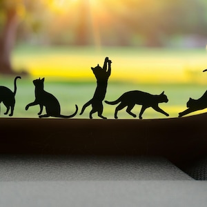 Peut inclure: Silhouette noire de cinq chats dans différentes poses, de debout à l'étirement. Les chats sont disposés en ligne sur un fond flou de verdure et de soleil. Le thème général est celui des chats.