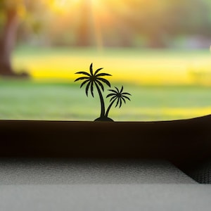 May include: Silhouette of two palm trees against a blurred background of green grass and a bright yellow sky. The palm trees are black and appear to be cut out of a solid material. The scene evokes a tropical or vacation theme.
