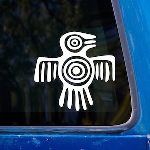 Puede incluir: Calcomanía de pájaro tribal blanco con un diseño estilizado, con círculos concéntricos y líneas verticales. La calcomanía está adherida a una ventana, sobre un fondo borroso y un marco de vehículo azul.