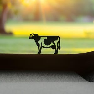 Könnte beinhalten: Ein schwarzer Kuh-Silhouetten-Aufkleber mit einem stilisierten Design mit weißen Negativraum-Flecken, der auf einer flachen Oberfläche angebracht ist. Die Grafik zeigt das Seitenprofil einer Kuh mit vier Beinen, einem Schwanz und einem Euter.