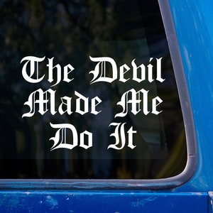 Puede incluir: Calcomanía de vinilo blanco con el texto "The Devil Made Me Do It" en un estilo de fuente gótico. La calcomanía está en una ventana de automóvil azul.