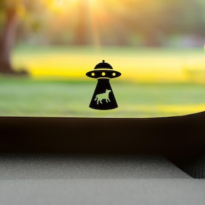 Può includere: Silhouette nera di un UFO con una mucca all'interno. L'UFO ha una classica forma a disco volante con luci. Lo sfondo è una scena sfocata all'aperto con erba verde e alberi e un cielo giallo brillante.